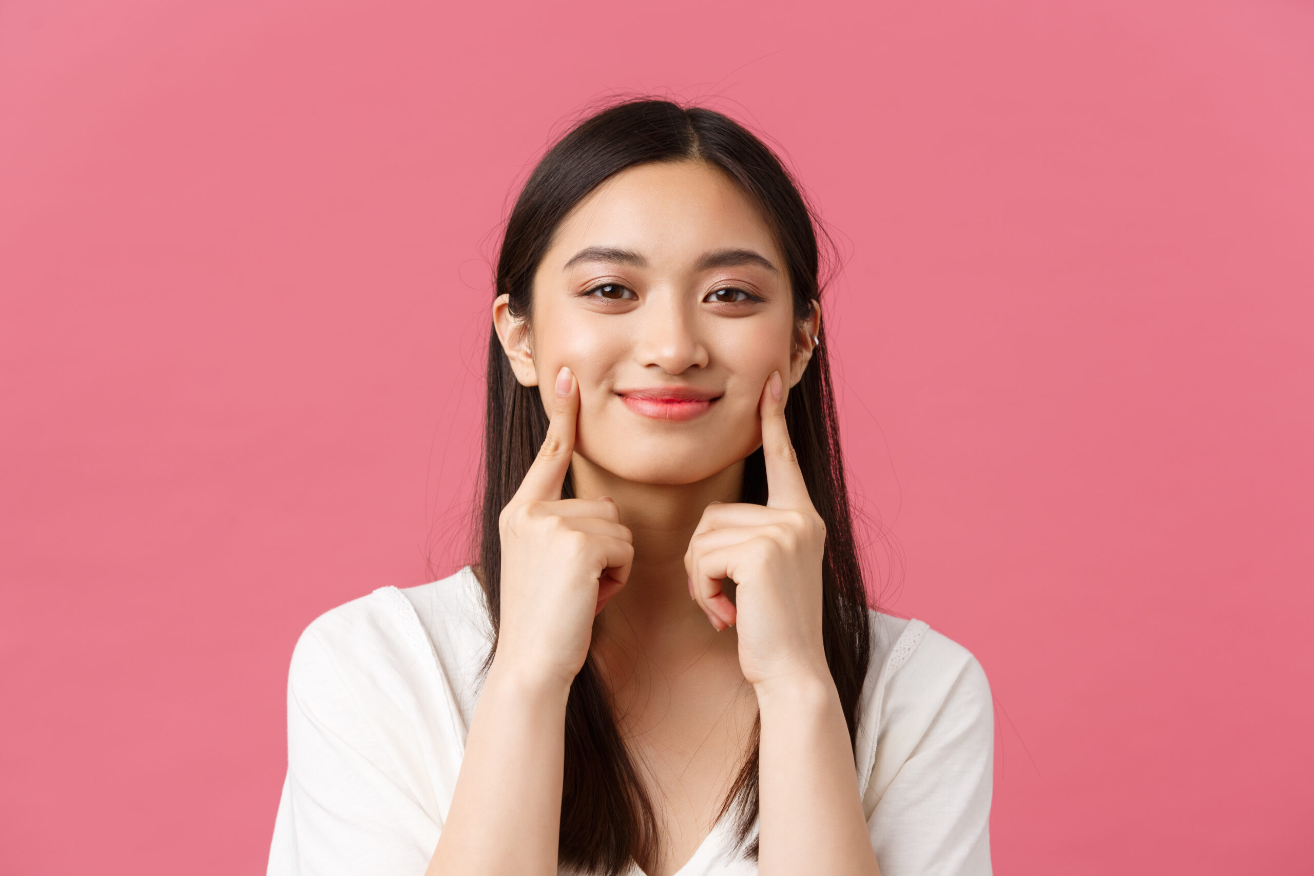 Beauty, people emotions and summer leisure concept. Close-up of funny and cute asian woman with kawaii dimples, touching cheeks and smiling happy, standing pink background